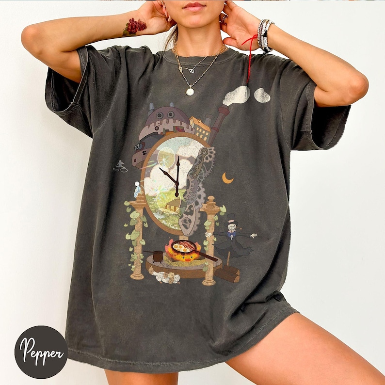 Op de afbeelding: Donkergrijs t-shirt met een fantasie klokontwerp met tandwielen, een landschap en cartoonfiguren. Het ontwerp omvat een wijzerplaat, een gebouw en een figuur in een pak. Het shirt heeft korte mouwen en een relaxte pasvorm.
