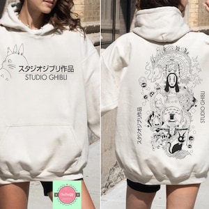 May include: Cream-colored Studio Ghibli hoodie with a front pocket and drawstring hood. The front features a black illustration of Totoro and Japanese text. The back displays a detailed black and white graphic of characters and Japanese text.