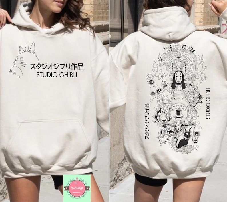 May include: Cream-colored Studio Ghibli hoodie with a front pocket and drawstring hood. The front features a black illustration of Totoro and Japanese text. The back displays a detailed black and white graphic of characters and Japanese text.