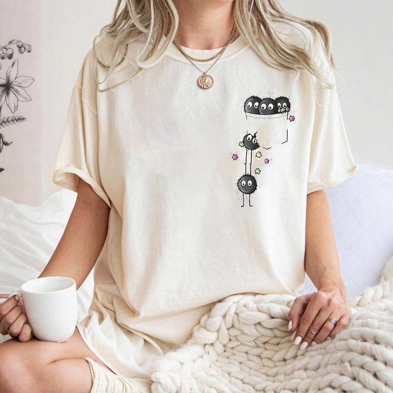 May include: Cream-colored t-shirt featuring a pocket design with cartoon soot sprites. The pocket contains three soot sprites, while two more are below, with colorful star accents. The shirt has short sleeves and a relaxed fit.