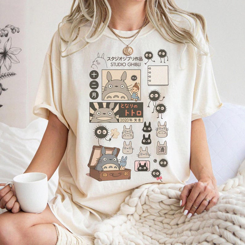 May include: Cream-colored t-shirt featuring a Studio Ghibli design with Japanese text and illustrations of Totoro and other characters. The graphic includes a suitcase, various character sketches, and the text "2020".