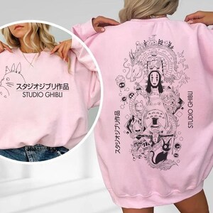May include: A light pink Studio Ghibli sweatshirt. The front features a Totoro graphic and Japanese text. The back displays a detailed black and white illustration of characters from Studio Ghibli films, with the words "Studio Ghibli" in English and Japanese.