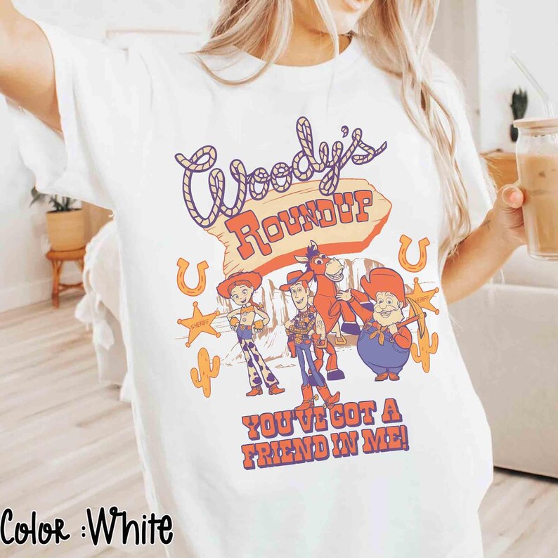 May include: White t-shirt featuring a colorful graphic of characters and text from "Woody's Roundup". The design includes the text "Woody's Roundup" and "You've Got a Friend in Me!" with images of cartoon characters, horseshoes, and cacti.
