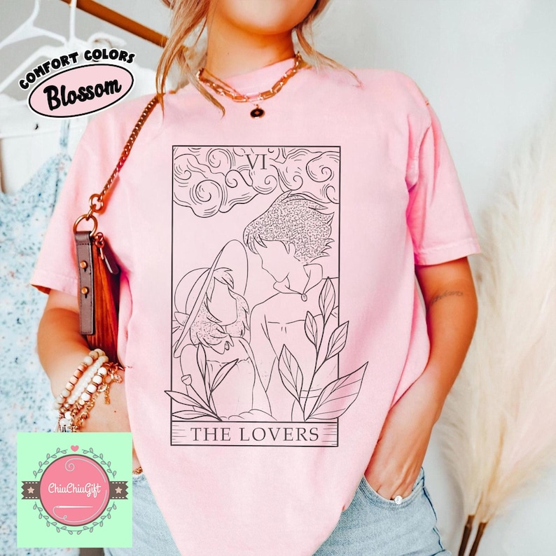 Puede incluir: Una camiseta rosa claro con una ilustraci&oacute;n en blanco y negro de la carta del tarot de Los Amantes. La camiseta tiene el texto "Comfort Colors Blossom" en la parte superior izquierda. La parte inferior de la ilustraci&oacute;n tiene el texto "THE LOVERS".