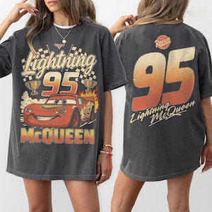 May include: A charcoal gray oversized t-shirt featuring a graphic of the red race car Lightning McQueen with the number 95, checkered flags, and trophies on the front, and a large 95 with the text Lightning McQueen on the back.