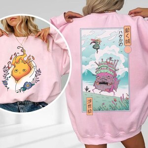 K&ouml;nnte beinhalten: Hellrosa Sweatshirt mit einem Grafik eines Feuergeistes in einer Hand auf der Vorderseite. Die R&uuml;ckseite zeigt eine japanische Anime-Szene mit einer Burg, einer Figur und Text. Das Sweatshirt ist aus weichem Material.