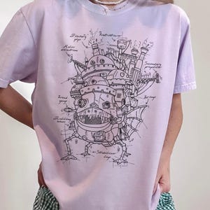 May include: A light purple t-shirt featuring a detailed black and white illustration of a mechanical structure with various labeled components. The design includes text such as "Discharge pipe" and "Wind gauge".
