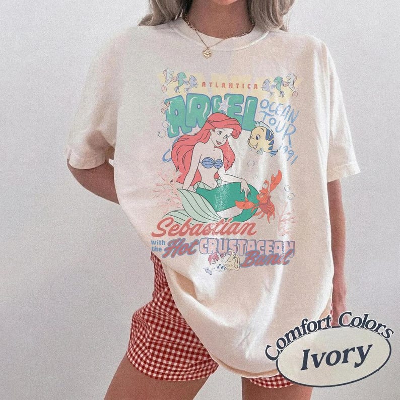 Op de afbeelding: Een ivoorkleurig T-shirt met korte mouwen en een grafische print van Ari&euml;l, Botje en Sebastiaan. De tekst luidt Atlantica Ocean Tour 1991, Sebastian with the Hot Crustacean Band. Het T-shirt heeft een ronde hals en een losse pasvorm.