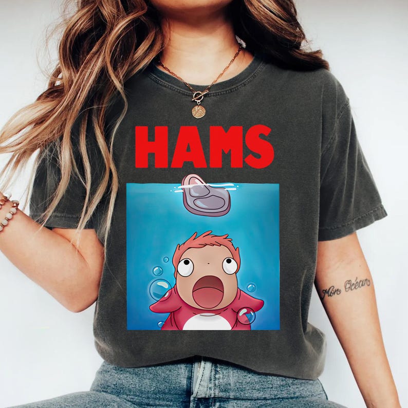 May include: Dark grey t-shirt featuring the word "HAMS" in red. Below is a cartoon illustration of a character looking upwards at a piece of meat in water. The character is red and white.