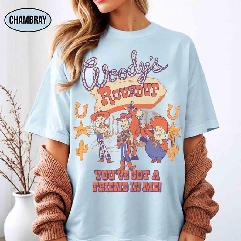 May include: Light blue t-shirt featuring the text "Woody's Roundup" and cartoon characters. The shirt also includes the phrase "You've Got A Friend In Me!" in orange and red lettering. The design includes a horseshoe, star, and cactus.