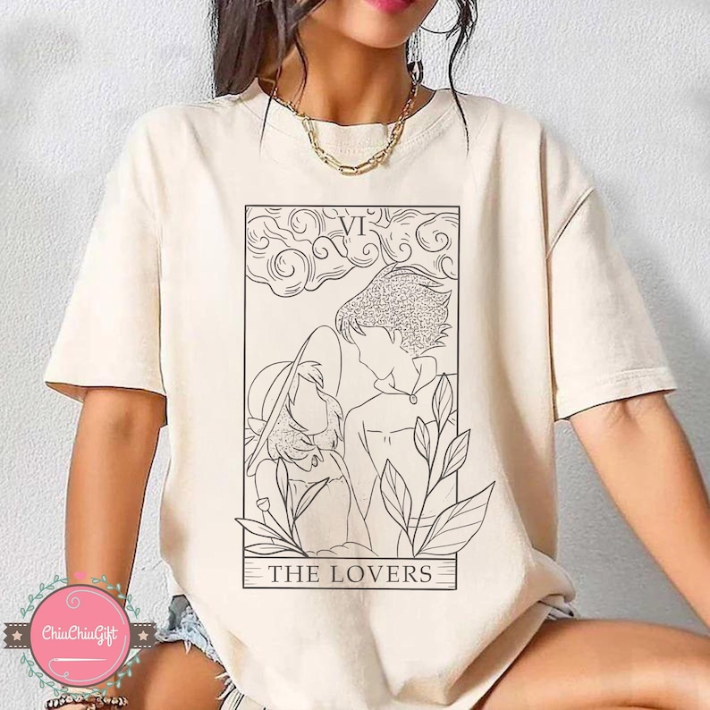 Puede incluir: Camiseta color crema con una ilustraci&oacute;n en negro de una carta del tarot. La carta representa dos figuras, con el texto "THE LOVERS" en la parte inferior. La parte superior de la carta tiene el n&uacute;mero romano "VI".