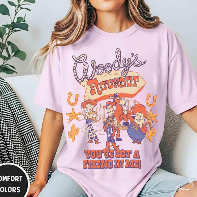 May include: A light purple t-shirt featuring the text "Woody's Roundup" in a retro font, with cartoon characters and the phrase "You've Got a Friend in Me!" printed below. The design includes a horseshoe, cactus, and sheriff's badge.