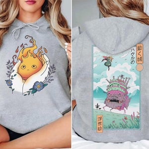K&ouml;nnte beinhalten: Ein hellgrauer Kapuzenpullover mit einem Frontmotiv eines Cartoon-Feuergeistes, der in H&auml;nden gehalten wird, umgeben von Blumen und Bl&auml;ttern. Das R&uuml;ckmotiv zeigt ein Schloss mit japanischem Text und eine Figur, die am Himmel fliegt.