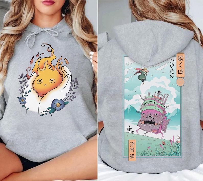 K&ouml;nnte beinhalten: Ein hellgrauer Kapuzenpullover mit einem Frontmotiv eines Cartoon-Feuergeistes, der in H&auml;nden gehalten wird, umgeben von Blumen und Bl&auml;ttern. Das R&uuml;ckmotiv zeigt ein Schloss mit japanischem Text und eine Figur, die am Himmel fliegt.