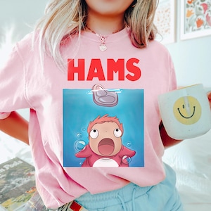 May include: Pale pink sweatshirt with the word "HAMS" in red. The graphic shows a cartoon character in blue water with a floating object. A white mug with a yellow smiley face is held in the image.