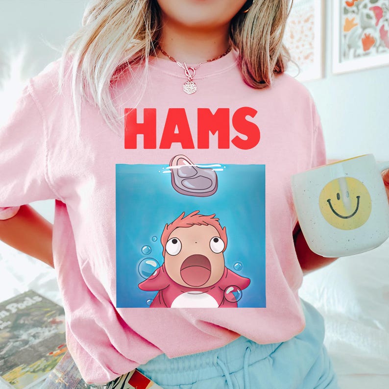 May include: Pale pink sweatshirt with the word "HAMS" in red. The graphic shows a cartoon character in blue water with a floating object. A white mug with a yellow smiley face is held in the image.