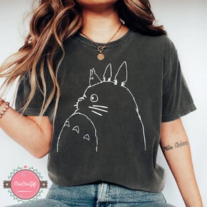 Puede incluir: Camiseta gris oscuro con un contorno blanco de Totoro y un personaje m&aacute;s peque&ntilde;o. El gr&aacute;fico est&aacute; centrado en la camiseta. La camiseta es de manga corta y cuello redondo.
