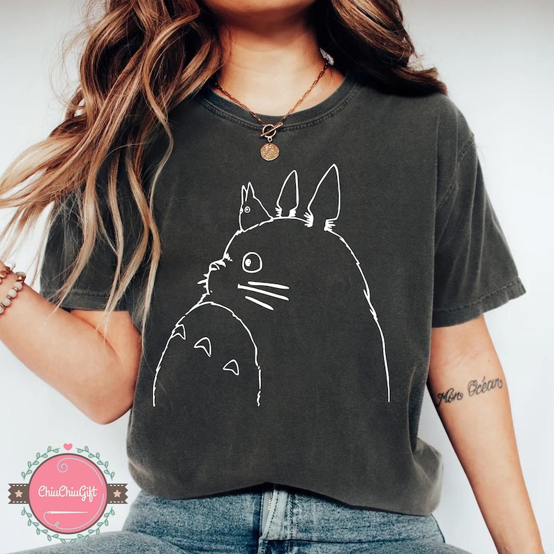 Puede incluir: Camiseta gris oscuro con un contorno blanco de Totoro y un personaje m&aacute;s peque&ntilde;o. El gr&aacute;fico est&aacute; centrado en la camiseta. La camiseta es de manga corta y cuello redondo.