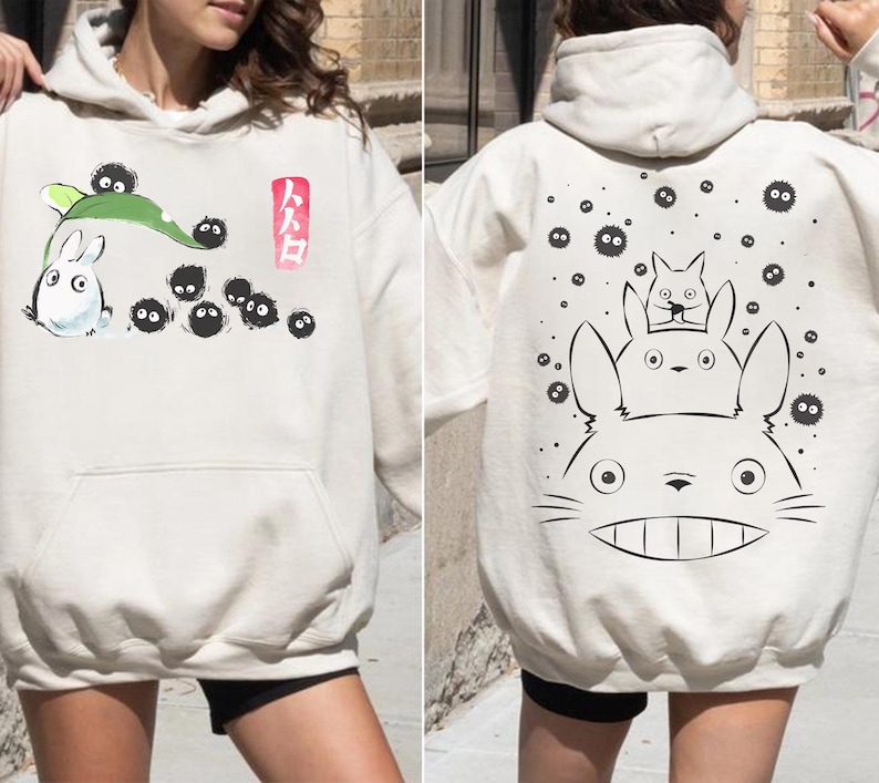 May include: Cream-colored hoodie featuring illustrations from a Japanese animated film. The front displays a white rabbit-like creature with black soot sprites and Japanese text. The back shows a large character with smaller characters.
