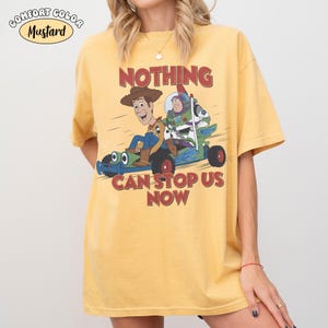 May include: A mustard yellow short-sleeve t-shirt featuring a graphic of Woody and Buzz Lightyear riding a green race car. The text above reads NOTHING and below reads CAN STOP US NOW. A small oval label in the corner says COMFORT COLOR MUSTARD.
