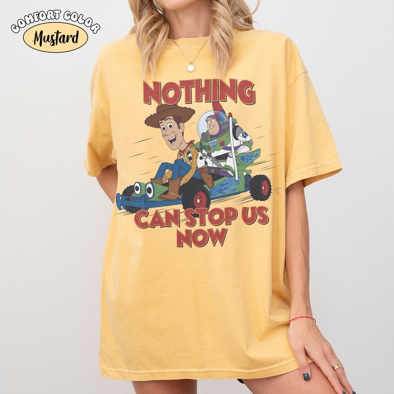 May include: A mustard yellow short-sleeve t-shirt featuring a graphic of Woody and Buzz Lightyear riding a green race car. The text above reads NOTHING and below reads CAN STOP US NOW. A small oval label in the corner says COMFORT COLOR MUSTARD.