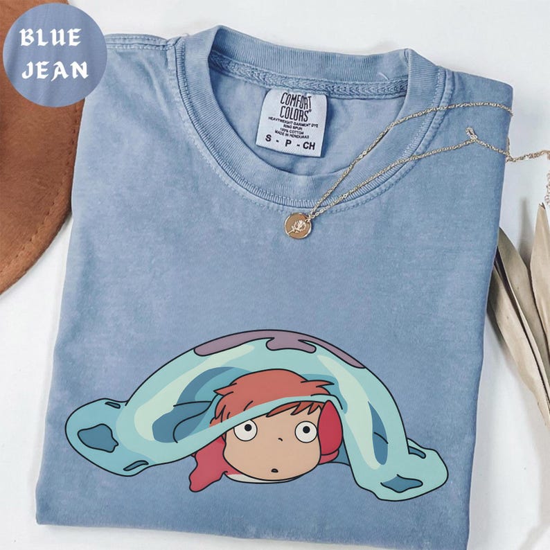 May include: Blue jean colored t-shirt with a cartoon graphic of a character under a blanket. The shirt has a crew neck and short sleeves. A gold necklace with a pendant is draped across the shirt. The tag reads "Comfort Colors".