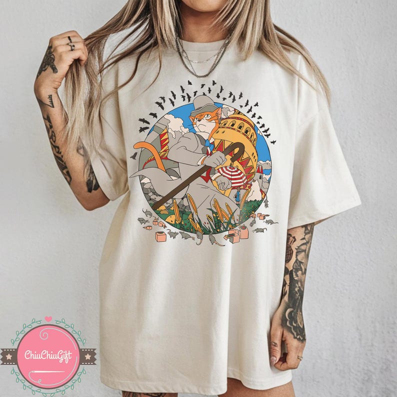 May include: Cream-colored t-shirt featuring a whimsical illustration of a cat dressed in a suit and hat, holding a cane. The cat is set against a backdrop of a building and flying bats. The shirt has a round neck and short sleeves.