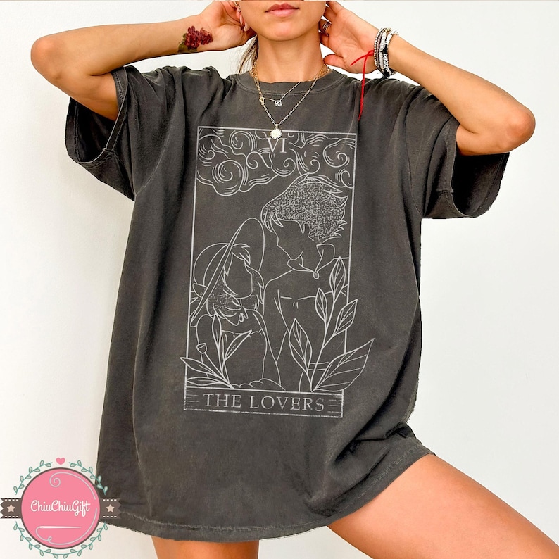 Puede incluir: Camiseta gris oscuro de gran tama&ntilde;o con un dibujo lineal blanco de la carta del tarot de Los Amantes. El dise&ntilde;o incluye dos figuras, follaje y el texto "THE LOVERS". La camiseta tiene mangas cortas y un corte holgado.