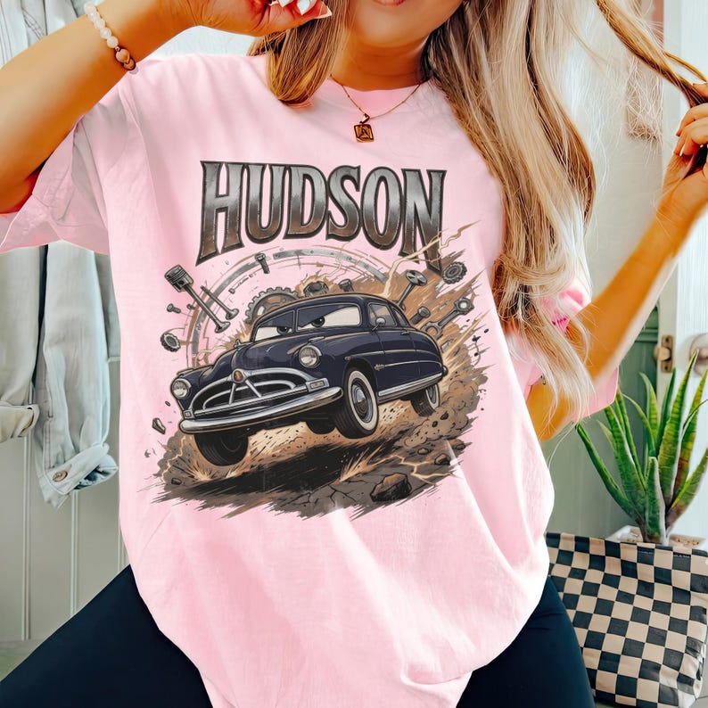 Pode incluir: Uma t-shirt rosa claro com uma estampa gr&aacute;fica de um carro antigo azul escuro com um rosto expressivo. A palavra HUDSON est&aacute; impressa em letras grandes, ousadas e de tom met&aacute;lico acima do carro, rodeada por pe&ccedil;as mec&acirc;nicas como engrenagens.