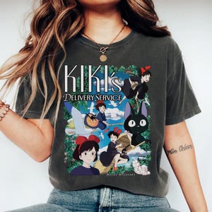 May include: Dark gray t-shirt featuring the text "KIKI's DELIVERY SERVICE" above a colorful graphic. The graphic depicts characters from the animated film, including a girl with a red bow, a black cat, and a flying broom.
