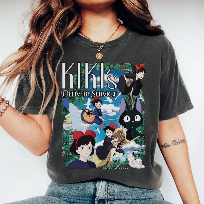 May include: Dark gray t-shirt featuring the text "KIKI's DELIVERY SERVICE" above a colorful graphic. The graphic depicts characters from the animated film, including a girl with a red bow, a black cat, and a flying broom.