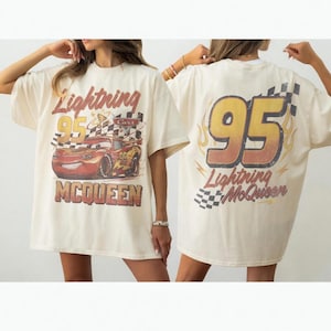 May include: An off-white t-shirt featuring a graphic of the red race car Lightning McQueen with the text Lightning McQueen and 95 on the front. The back displays a large 95 with flame accents, checkered patterns, and the text Lightning McQueen.