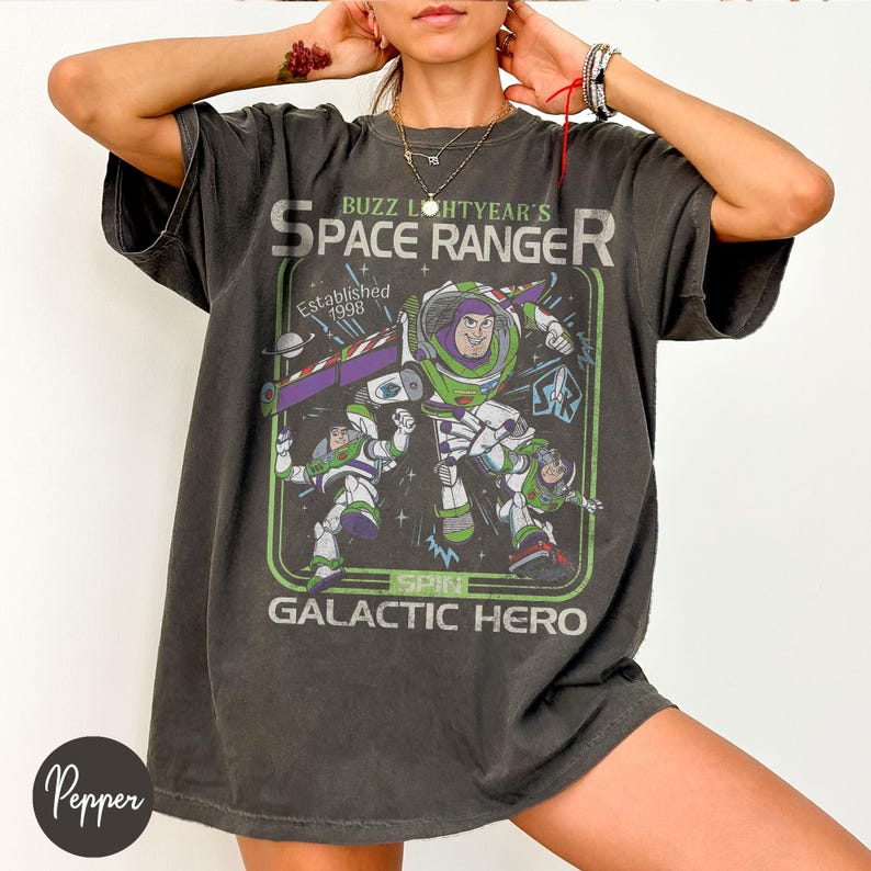 May include: Dark grey t-shirt with a graphic of Buzz Lightyear, a space ranger, and the text "Buzz Lightyear's Space Ranger" and "Galactic Hero". The design includes a green and purple spaceship and the year "Established 1998".