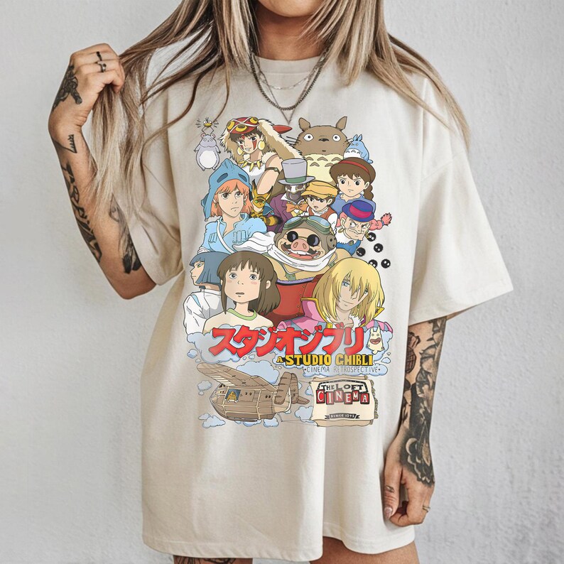 May include: Cream-colored t-shirt featuring a colorful graphic of Studio Ghibli characters. The design includes Japanese text and the words "THE LOFT CINEMA RETROSPECTIVE". The shirt has a relaxed fit and short sleeves.
