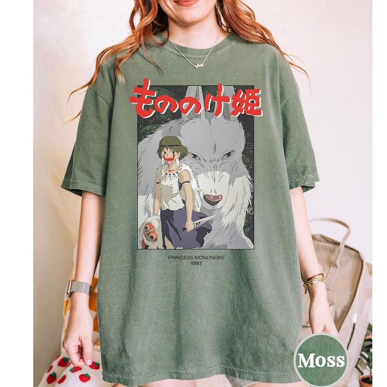May include: An olive green t-shirt with a graphic from the animated film "Princess Mononoke." The design includes Japanese text, a girl, and a wolf. The text "PRINCESS MONONOKE 1997" is also visible.