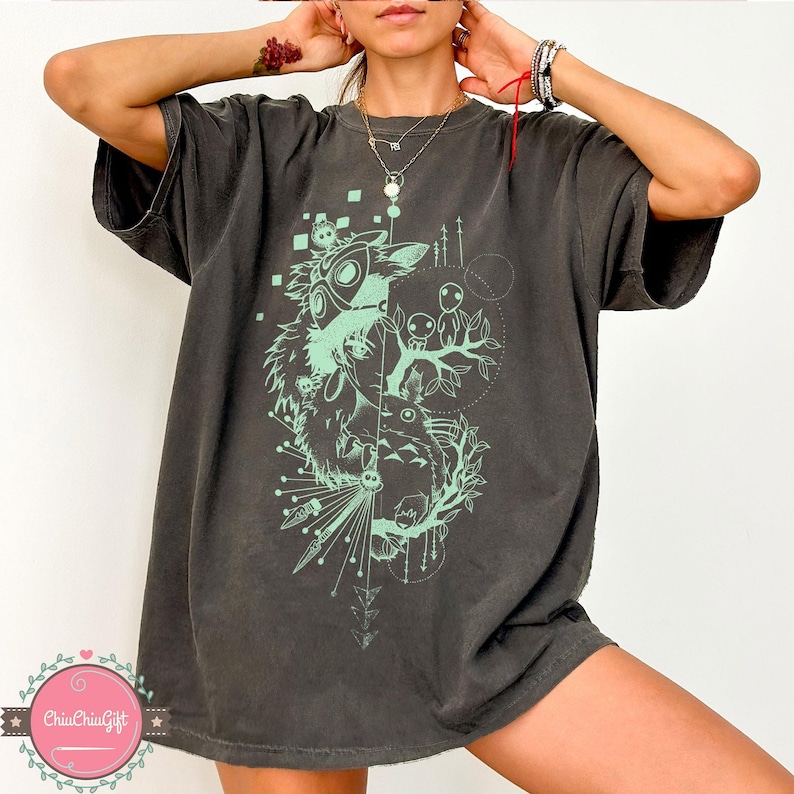 Puede incluir: Camiseta gris oscuro con un dise&ntilde;o gr&aacute;fico verde menta. El dise&ntilde;o incluye una ilustraci&oacute;n estilizada de un esp&iacute;ritu del bosque, &aacute;rboles y formas geom&eacute;tricas. La camiseta tiene un corte holgado y mangas cortas.