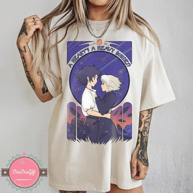 Puede incluir: Camiseta color crema con un gr&aacute;fico de dos personajes de anime abraz&aacute;ndose bajo un cielo estrellado. El texto "A Heart's A Heavy Burden" est&aacute; sobre la imagen. La camiseta tiene un corte holgado.