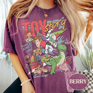 May include: A berry-colored t-shirt featuring a vintage-style Toy Story graphic. The print includes characters Jessie, Woody, Buzz Lightyear, Rex, Slinky Dog, Bullseye, and Hamm, with the text "TOY story," "SINCE 1995," and character names.