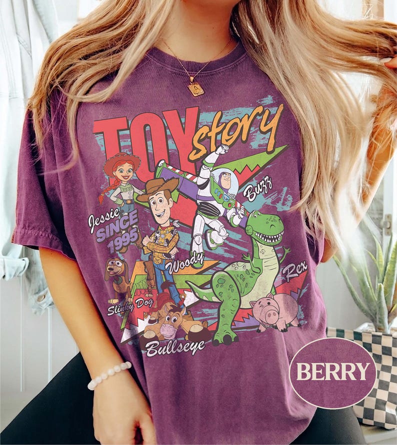 May include: A berry-colored t-shirt featuring a vintage-style Toy Story graphic. The print includes characters Jessie, Woody, Buzz Lightyear, Rex, Slinky Dog, Bullseye, and Hamm, with the text "TOY story," "SINCE 1995," and character names.