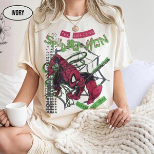May include: An ivory short-sleeve t-shirt featuring a graphic print of the comic book character Spider-Man in red and blue. The text reads THE AMAZING SPIDER-MAN and QUEENS NEW YORK. The design includes a black spiderweb pattern and geometric shapes.