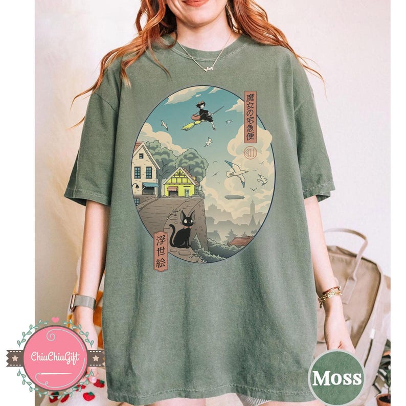Op de afbeelding: Mosgroen t-shirt met een cirkelvormige afbeelding ge&iuml;nspireerd op een Japanse animatiefilm. De afbeelding toont een heks op een bezem, een zwarte kat, huizen en vliegende vogels, met Japanse tekst.