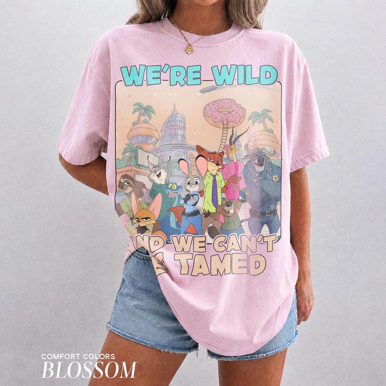 May include: A light pink short-sleeve t-shirt featuring a graphic print of various animated animal characters from Zootopia. The text WE'RE WILD AND WE CAN'T BE TAMED is printed in light blue and beige. The shirt has a crew neckline and a relaxed fit.