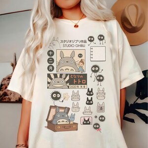 May include: Cream-colored t-shirt featuring Studio Ghibli characters and Japanese text. The design includes Totoro, soot sprites, and other illustrations. The shirt has a crew neck and short sleeves.