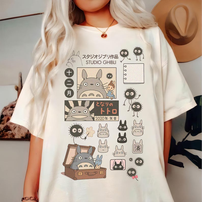 May include: Cream-colored t-shirt featuring Studio Ghibli characters and Japanese text. The design includes Totoro, soot sprites, and other illustrations. The shirt has a crew neck and short sleeves.