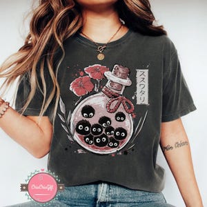 Puede incluir: Camiseta gris oscuro con una ilustraci&oacute;n de fantas&iacute;a. Presenta una botella de poci&oacute;n con duendes de holl&iacute;n negros, flores rojas y texto japon&eacute;s. La botella tiene un tap&oacute;n de corcho y est&aacute; atada con una cuerda roja.