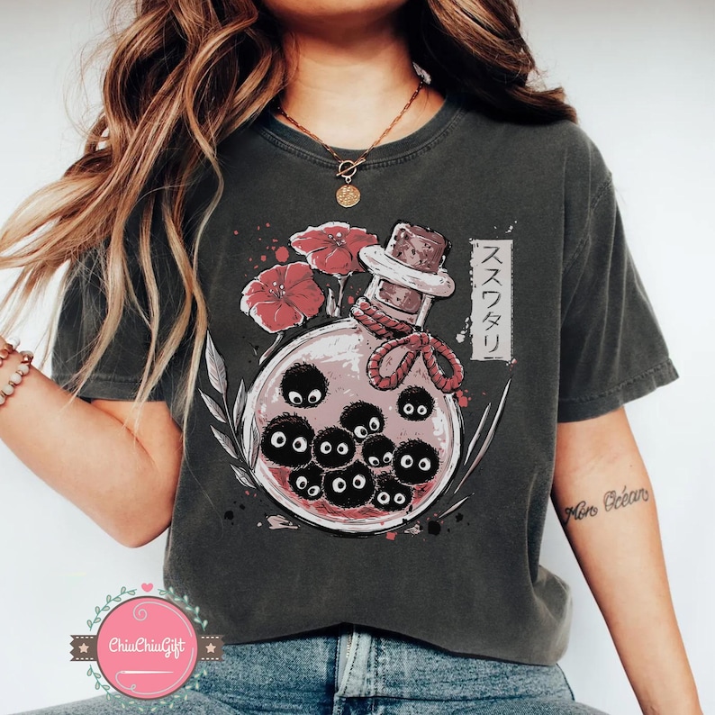 Puede incluir: Camiseta gris oscuro con una ilustraci&oacute;n de fantas&iacute;a. Presenta una botella de poci&oacute;n con duendes de holl&iacute;n negros, flores rojas y texto japon&eacute;s. La botella tiene un tap&oacute;n de corcho y est&aacute; atada con una cuerda roja.