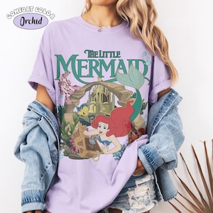 May include: An orchid-coloured t-shirt featuring a graphic print of The Little Mermaid with Ariel, Flounder, and Sebastian. The design includes an underwater castle, a treasure chest, and the text The Little Mermaid in green lettering.