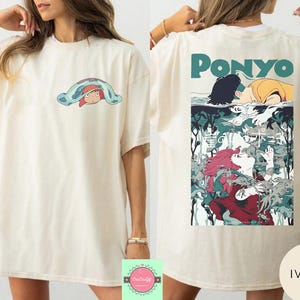 May include: Ivory-colored t-shirt featuring a cartoon graphic of a character under a blue blanket on the front. The back of the shirt displays a colorful illustration with the word "Ponyo" in teal.