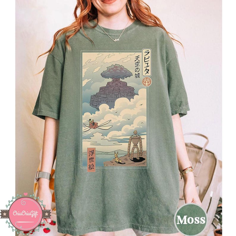 May include: Moss green t-shirt featuring a Japanese anime-style illustration. The design includes a floating castle, a robot, and characters in a sky scene. Japanese text is present in the design.