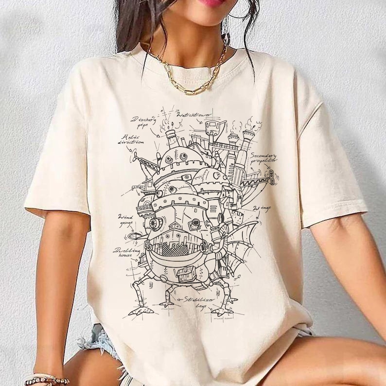 May include: Cream-colored t-shirt featuring a detailed black and white illustration of a fantastical, moving castle with architectural details and handwritten notes. The design includes text such as "Ducting pipe" and "Stabilizer legs".